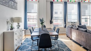 Een moderne woonkamer met een eetgedeelte, grote ramen met kleurrijk glas-in-lood, een grijze bank en een blauw, patroonrijk vloerkleed.