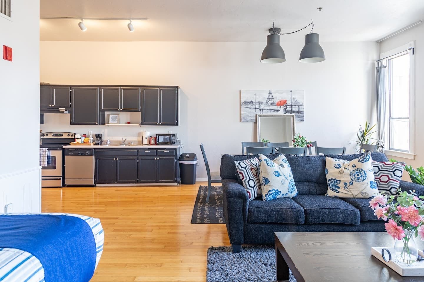 Eine moderne Küche mit dunklen Schränken, ein Essbereich mit grauem Sofa und ein Wohnzimmer mit blauem Sofa und Zierkissen.