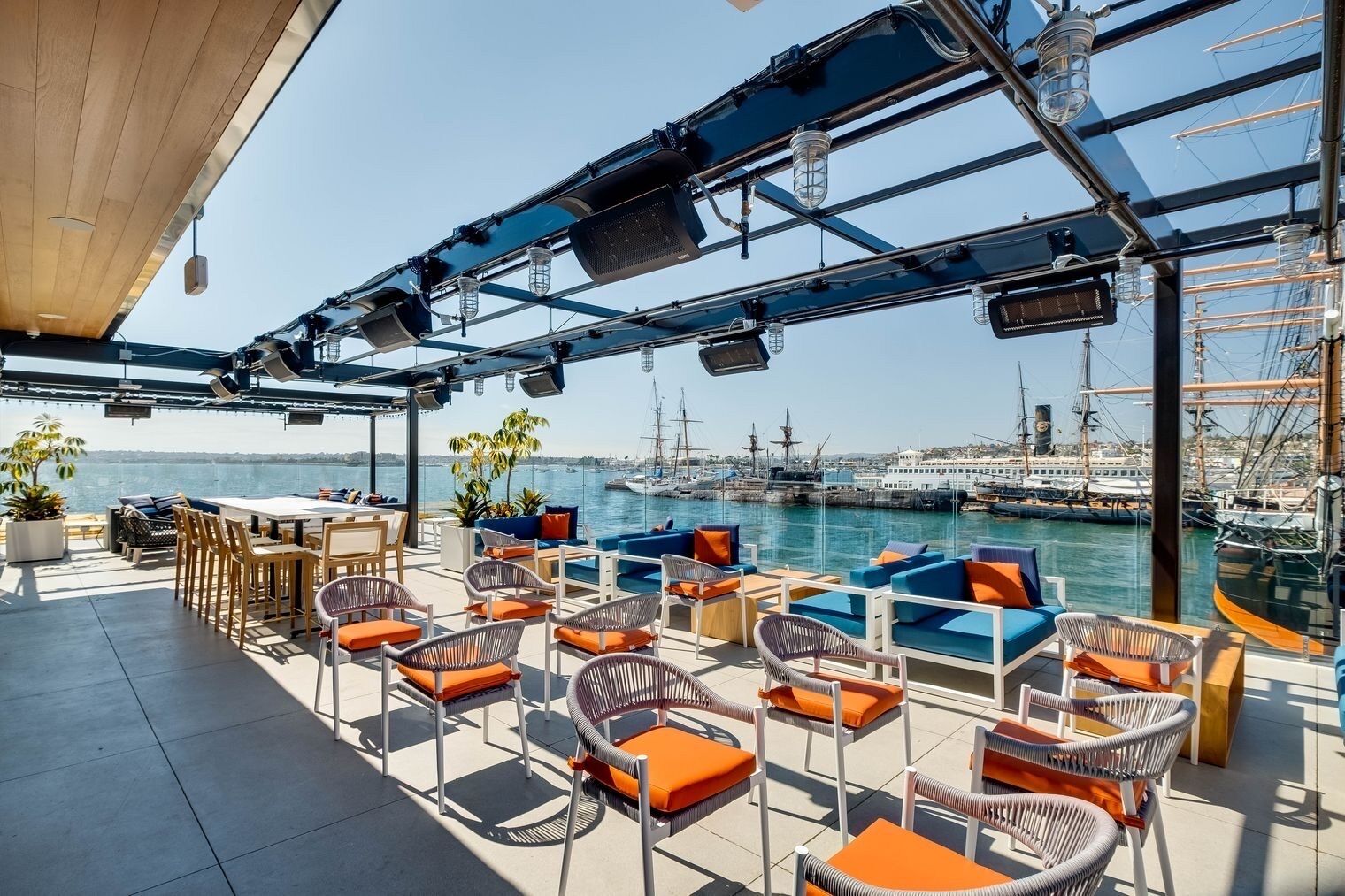A covered outdoor seating area with wicker chairs and cushions, overlooking a marina with boats and a clear sky.