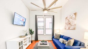 A living room with a blue sofa, a white entertainment unit, a ceiling fan, a TV, and a window with curtains.