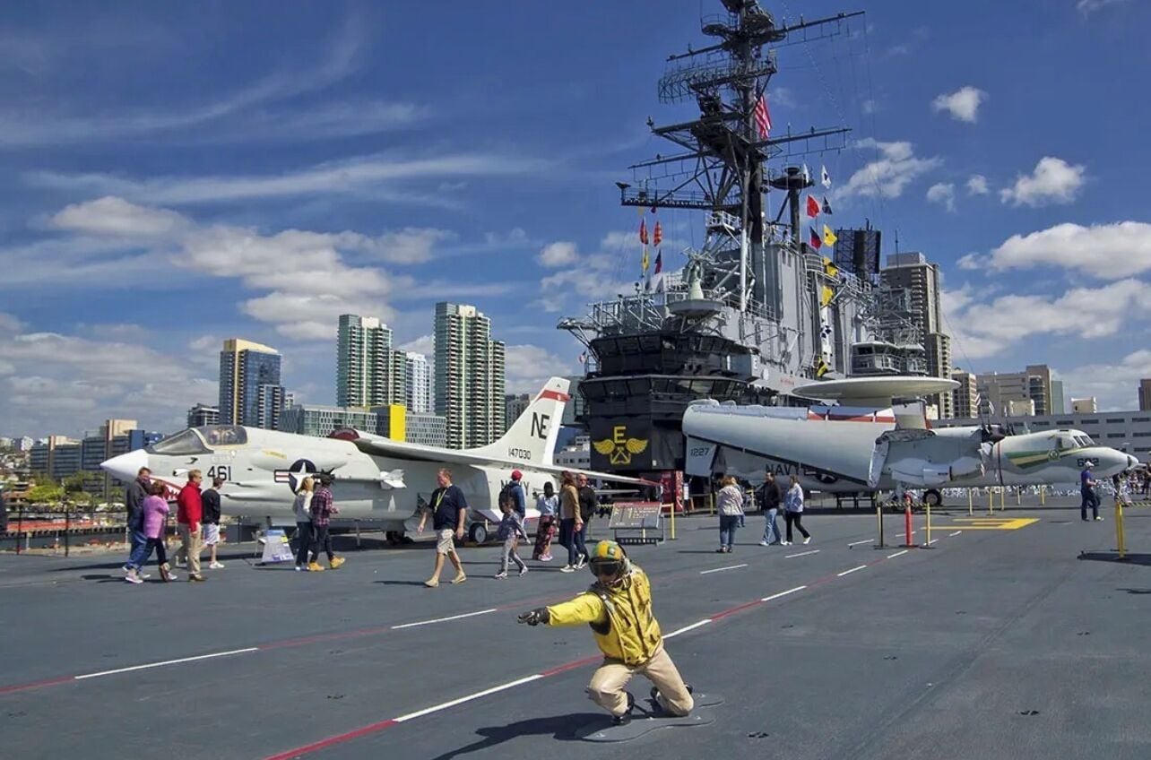 A naval aircraft carrier with multiple aircraft parked on its deck, surrounded by a city skyline.