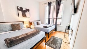 Ein modernes Schlafzimmer mit zwei Betten, einem großen Fenster mit Vorhängen, einem Wand-Fernseher und einem kleinen Tisch mit einer Pflanze.