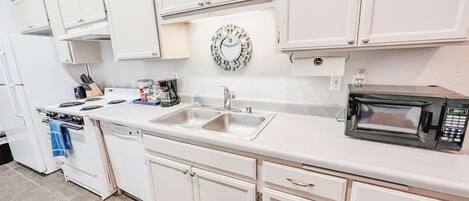 A kitchen with white cabinets, a stainless steel sink, a microwave, and a clock on the wall.