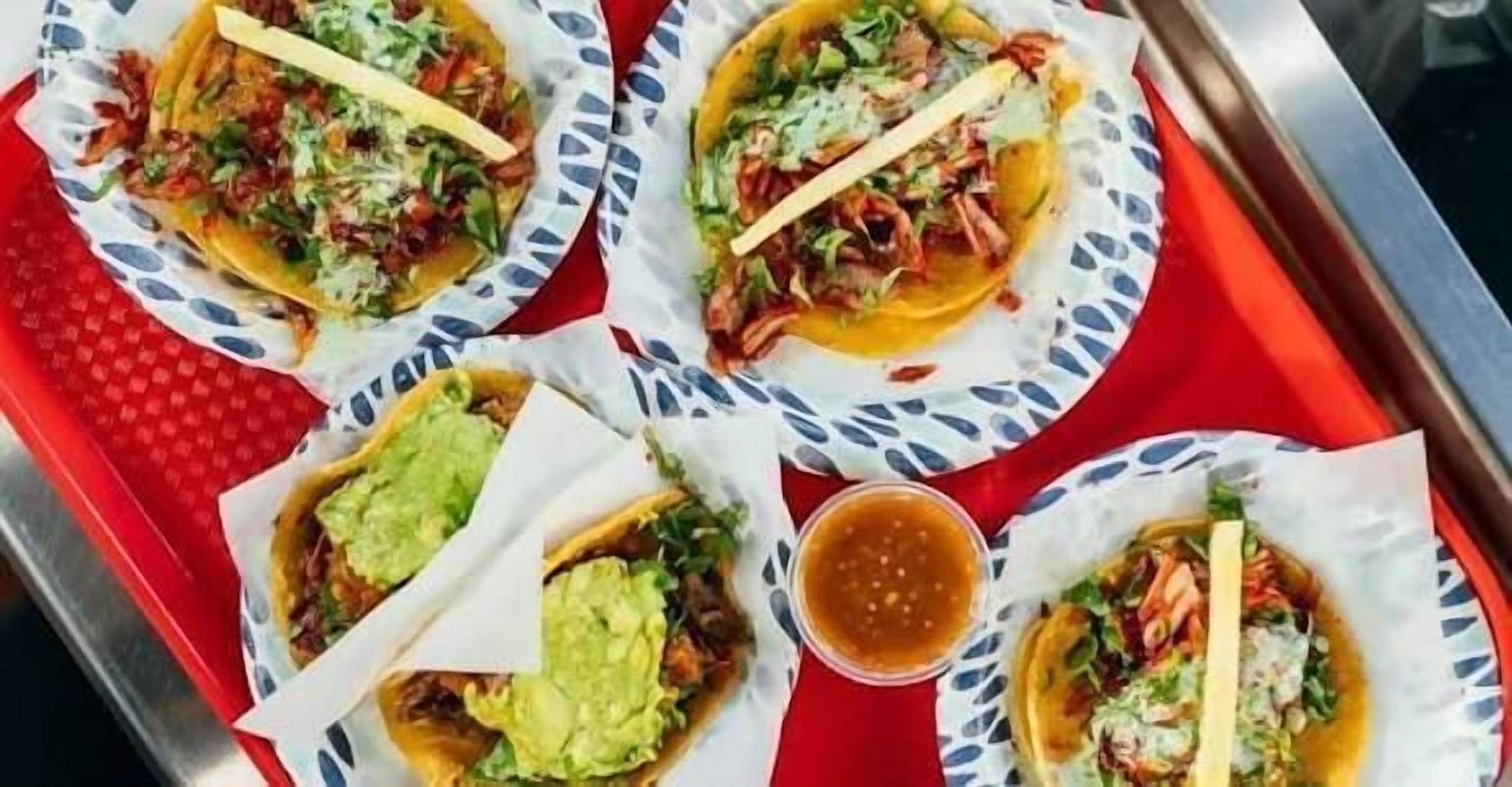 A tray with four tacos, each topped with different ingredients, and a small container of sauce.