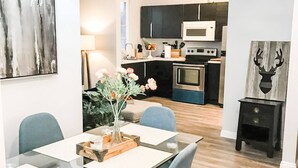 A modern dining area with a glass table, chairs, and a vase of flowers. A kitchen is visible in the background with dark cabinets and modern appliances.