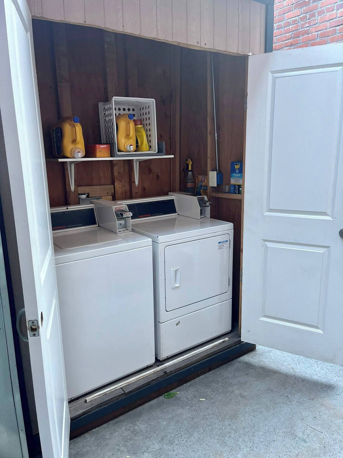 A laundry room with two front-loading washers and dryers, a shelf with cleaning supplies, and a door to the right.