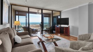 A spacious living room with a large sofa, a coffee table, and a TV. The room has a balcony with a view of the beach. - Beautiful 2BR/2BA ocean view condo that feels like home (Wrightsville Beach)