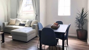 A modern living room with a grey sofa, a dining table with chairs, a potted plant, and a large window.