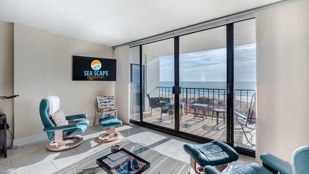 A balcony with a view of the beach, two teal chairs, a small table with a tray, and a TV displaying 'SEA SCAPE PROPERTIES'.