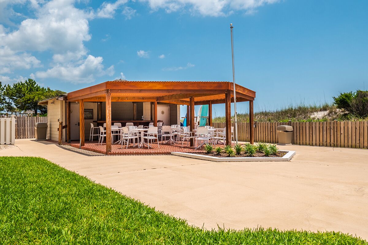 Un espacio de estar al aire libre cubierto, con mesas y sillas, un cerco de madera y un césped.