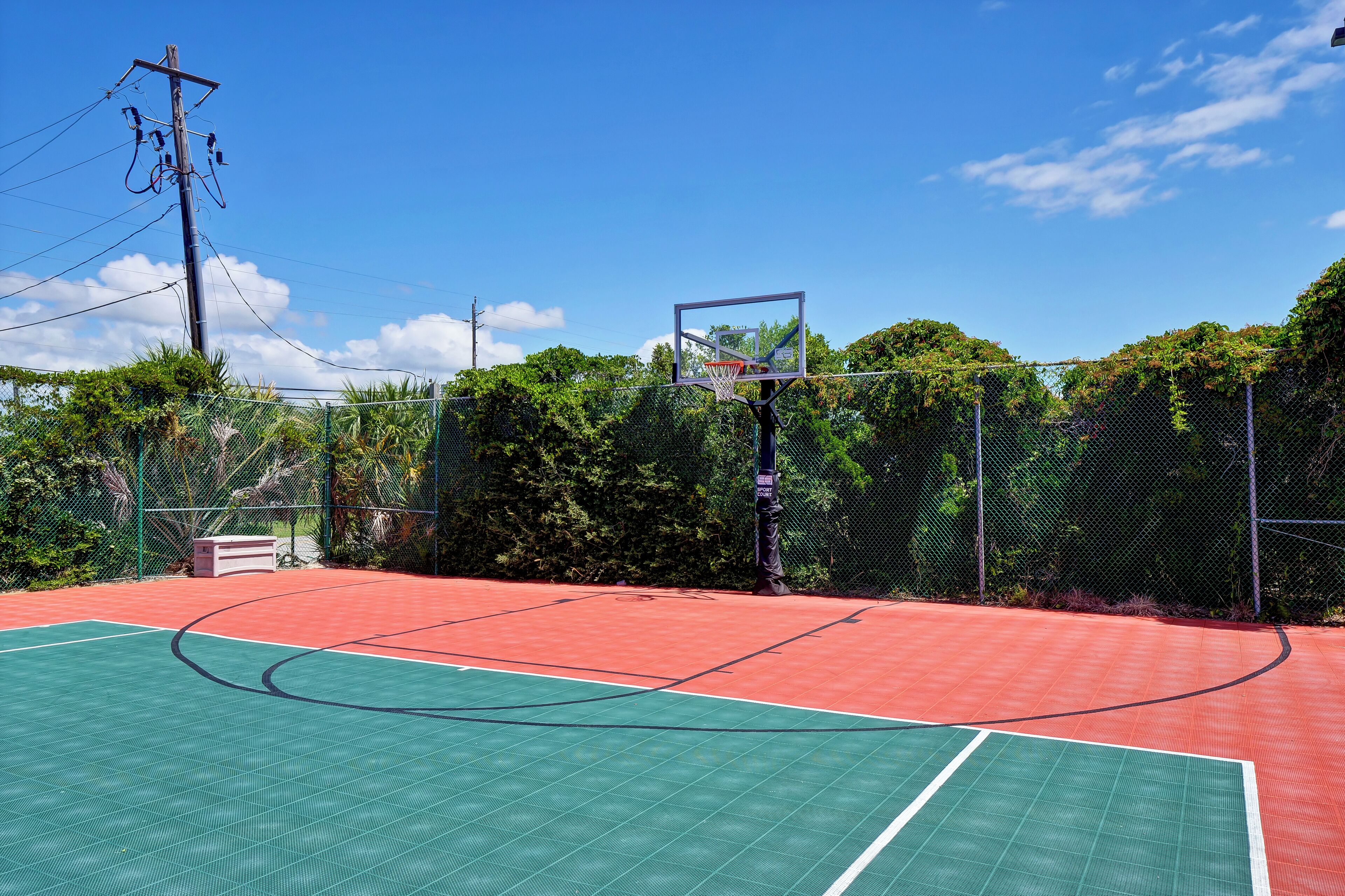 Een basketbalveld buiten met een rood en groen oppervlak, omgeven door een gaashek en begroeiing.