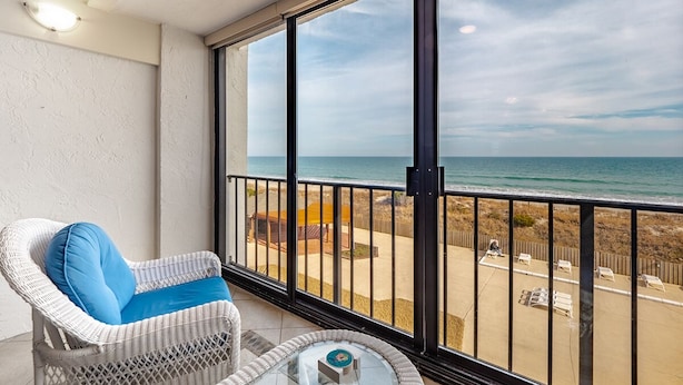 Ein Balkon mit Korbstuhl und Glastisch, der Blick auf einen Strand mit Zaun und Liegestühlen freigibt.