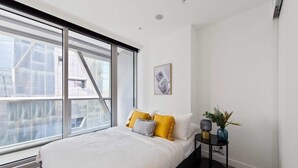 Un dormitorio con un ventanal grande, una cama con ropa de cama blanca y almohadas amarillas, una mesita de noche con una lámpara y un florero, y un cuadro en la pared.