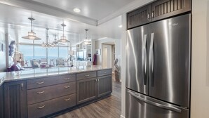 A modern kitchen with a large stainless steel refrigerator, dark cabinetry, and pendant lighting. - Modern 2BR/2BA condo unit w/ community pool and oceanfront view (Wrightsville Beach)