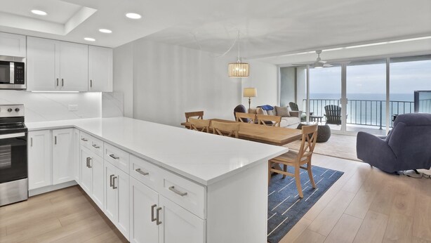 A modern kitchen with white cabinets, a central island, and a dining area with a wooden table and chairs. - Savor ocean views in this 2BR/2Ba condo w/community pool (Wrightsville Beach)