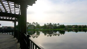 Una terraza de madera con barandilla da vista a un lago tranquilo, que refleja edificios y árboles cercanos.