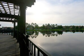 Eine Holzterrasse mit Geländer, die auf einen ruhigen See blickt, in dem nahegelegene Gebäude und Bäume sich spiegeln.