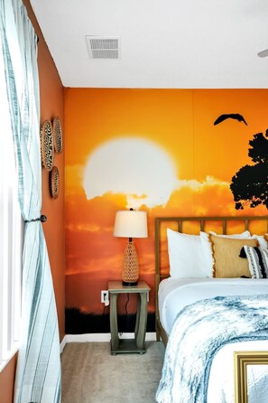 Un dormitorio con cama, mesita de noche, lámpara y un mural en la pared con un atardecer.