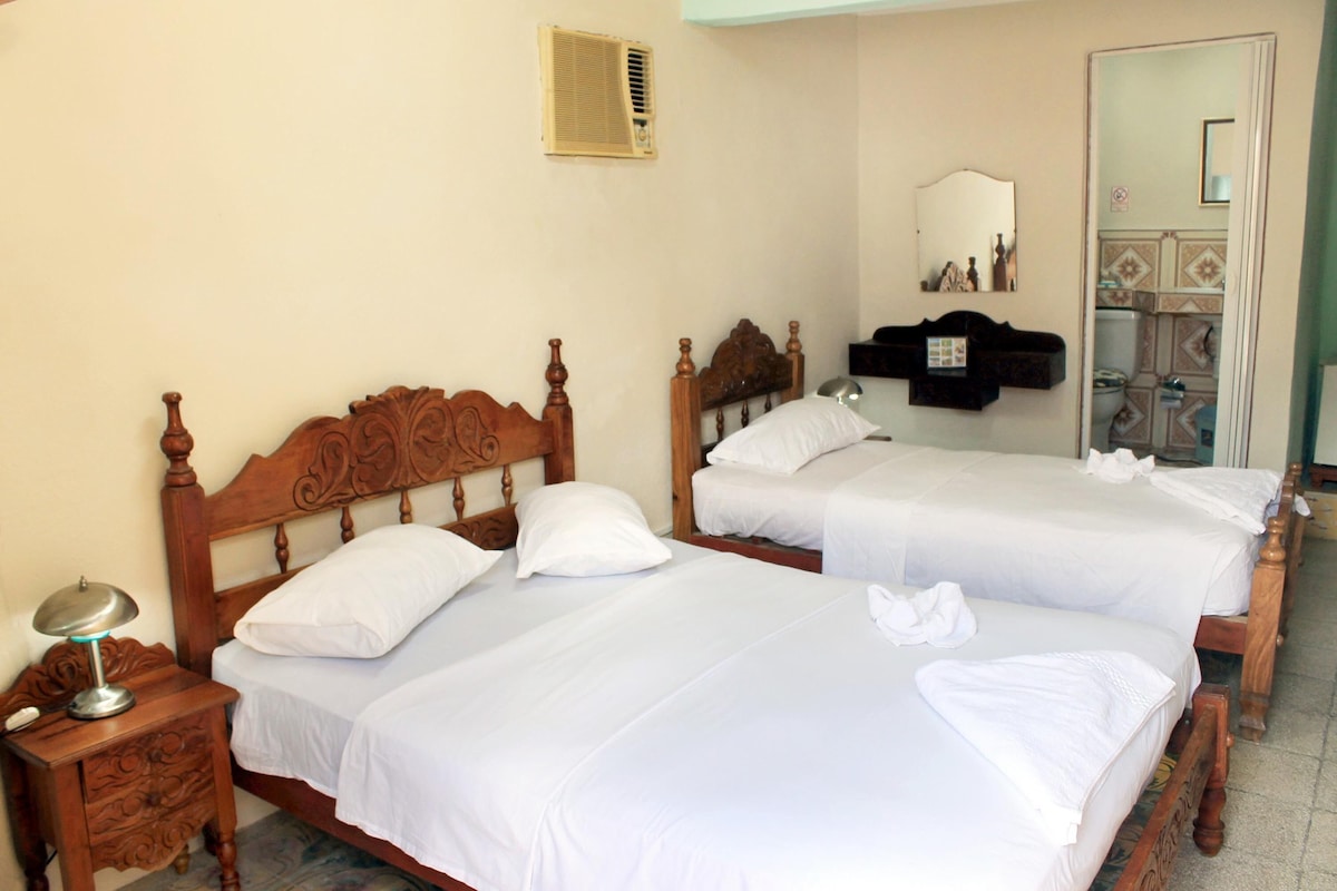 A hotel room with two wooden beds, white linens, a bedside table with a lamp, a mirror, and a shelf with toiletries.