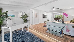 En rummelig opholdsstue med et sofabord i glas, en hvid sofa og et spisebord. Planter og en loftventilator er synlige.