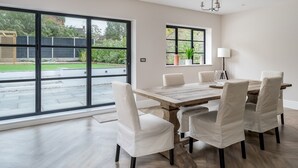 En matplats med ett stort bord och sex stolar, stora fönster med utsikt över en trädgård och byggnader, samt en modern kristallkrona.