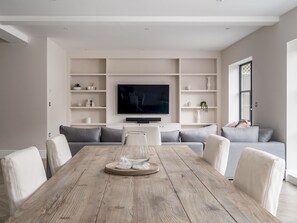 A modern living room with a wooden dining table, grey sofas, and built-in shelving.