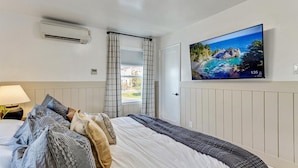 A bedroom with a bed, a TV on the wall, a window with curtains, and an air conditioning unit.