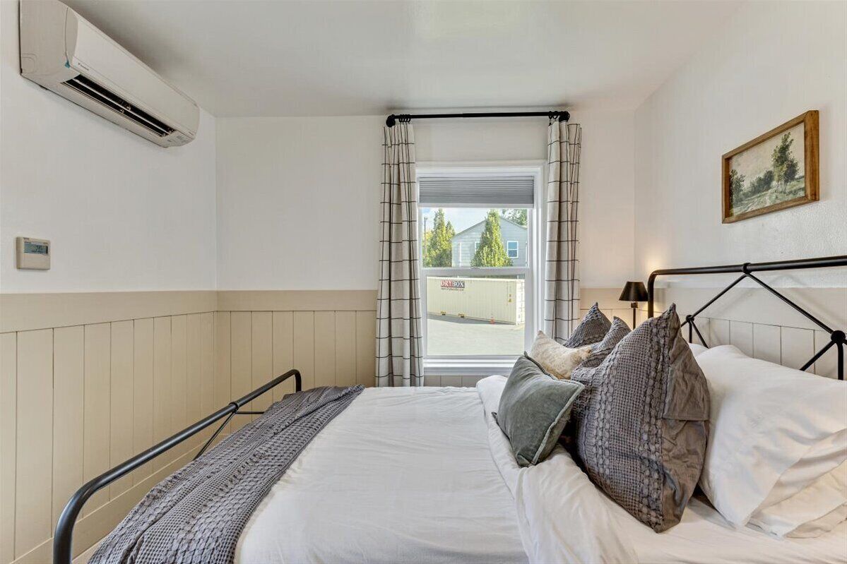 Un dormitorio con cama, ventana con cortinas, aire acondicionado y un cuadro en la pared.