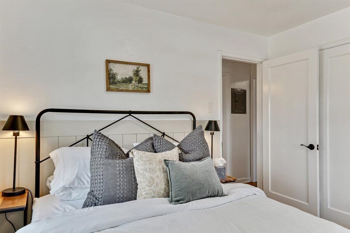 Una cama cuidadosamente tendida con estructura metálica negra, ropa de cama blanca y varias almohadas. Hay una lámpara de noche a cada lado de la cama, un cuadro enmarcado en la pared sobre la cama y una puerta a la derecha.