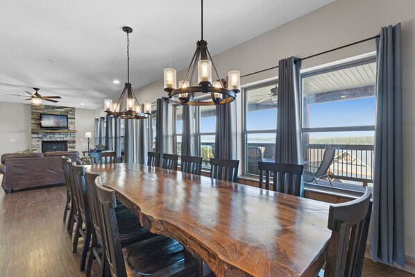 A spacious dining area with a long wooden table, multiple chairs, and a chandelier. There is a television mounted on a stone wall, and large windows with curtains. - Lakefront Haven, an Amazing 9 Bedroom Vacation Home Where the Fun Never Ends! (Branson)