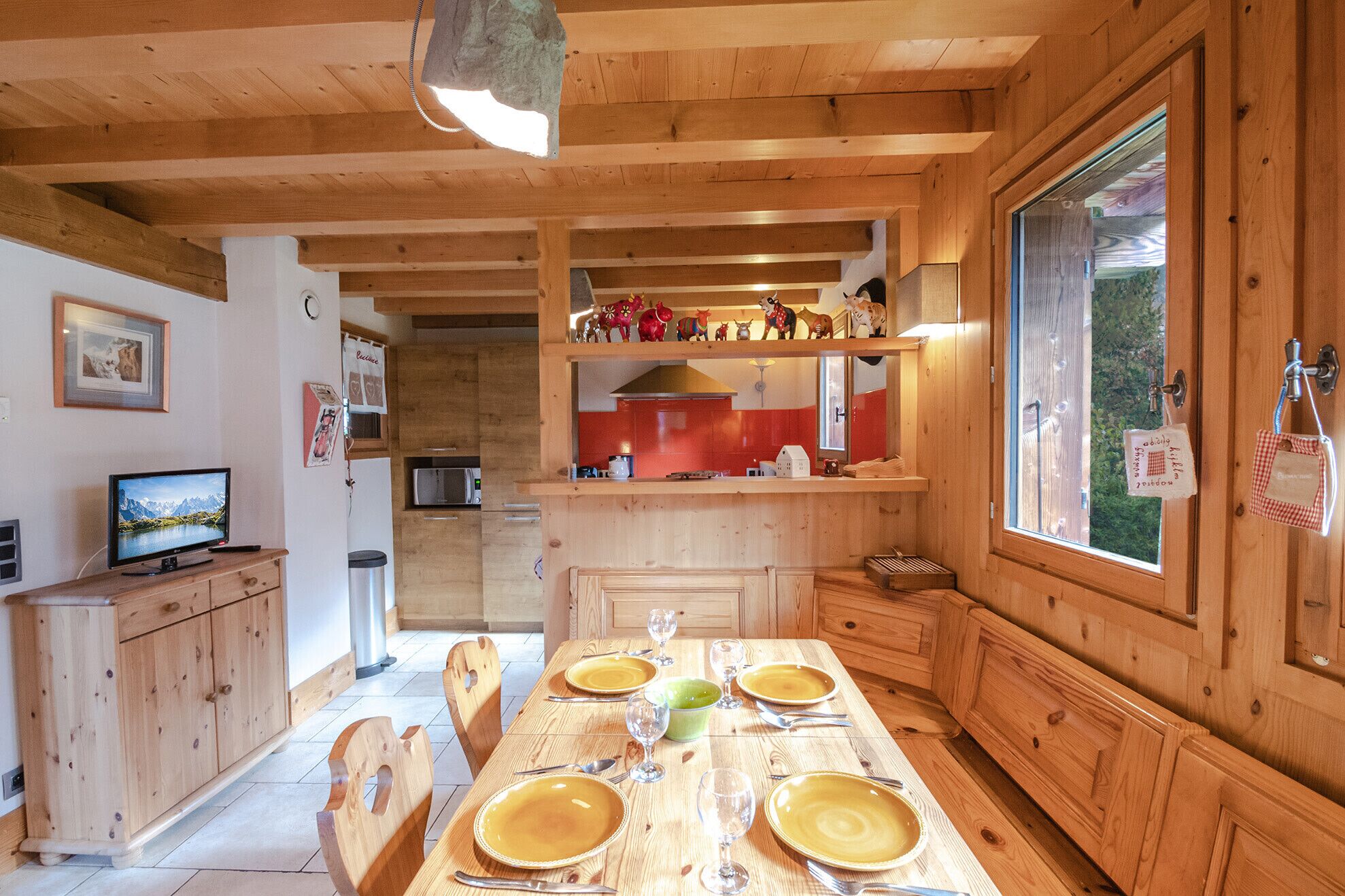 A wooden interior with a dining table set for four, a kitchen area with red cabinets, and a large window.