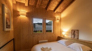 A wooden room with a bed, two windows, and a nightstand with a lamp. - Family chalet with terrace and garden in Chamonix (Chamonix-Mont-Blanc)