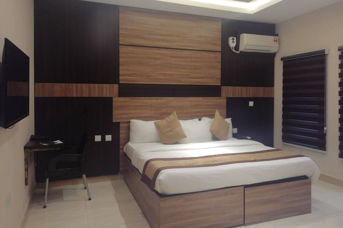 A modern bedroom with a wooden headboard, a flat-screen TV, an air conditioning unit, and a window with blinds.