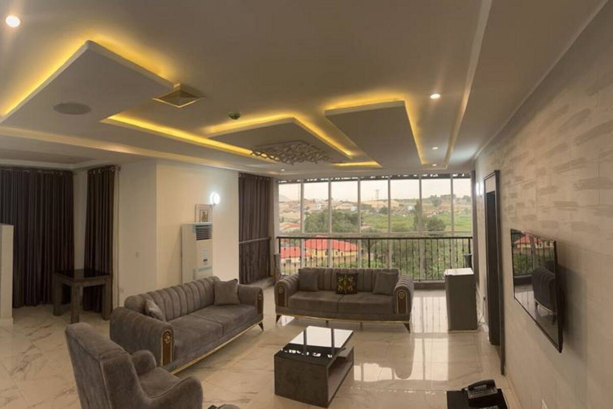 A modern living room with a sofa, a coffee table, and a TV. There is a kitchen area with cabinets and a window.