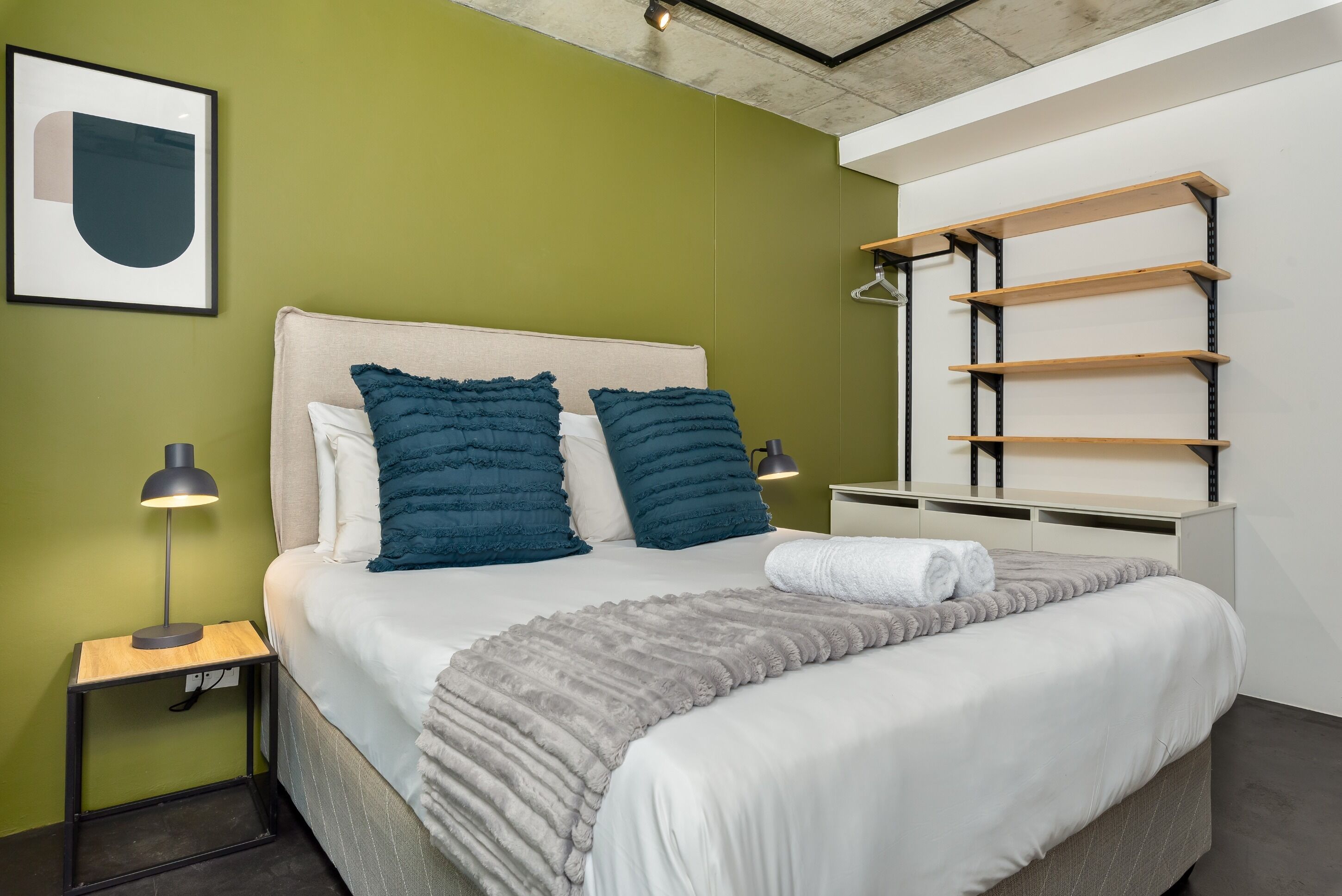 A neatly made bed with a grey blanket, two blue pillows, and a white pillow. There is a small bedside table with a lamp, and a wooden shelf unit against the wall.