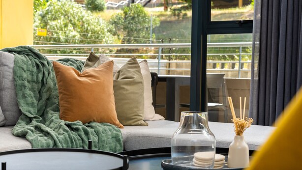 Ein modernes Wohnzimmer mit Sofa, Kissen, einem Glastisch und Blick auf Grünfläche.