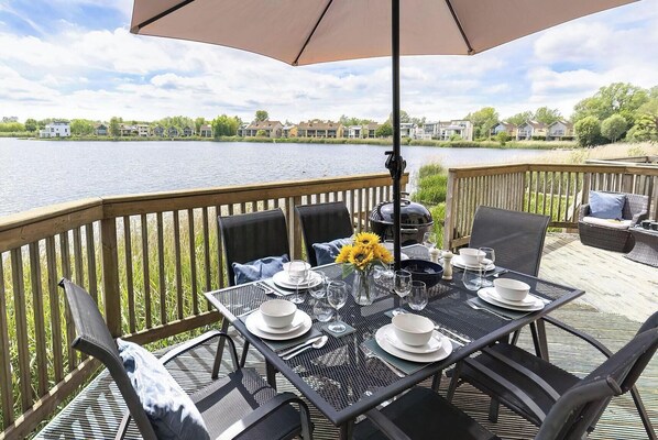 Une terrasse avec une table dressée pour le repas, donnant sur une étendue d’eau, avec des bâtiments en arrière-plan.
