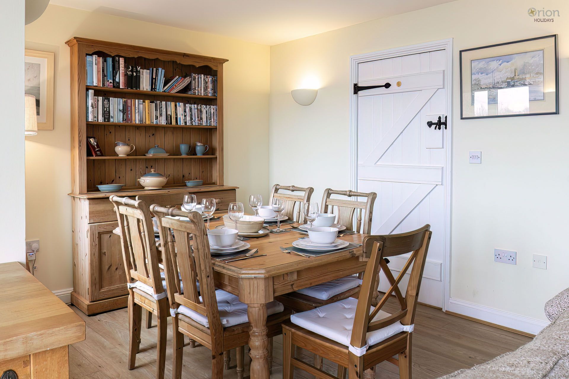 A dining room with a wooden table set for six, surrounded by six wooden chairs with cushions. A wooden bookshelf filled with books and decorative items is to the left. A white door with a black handle and a framed picture on the wall are visible.