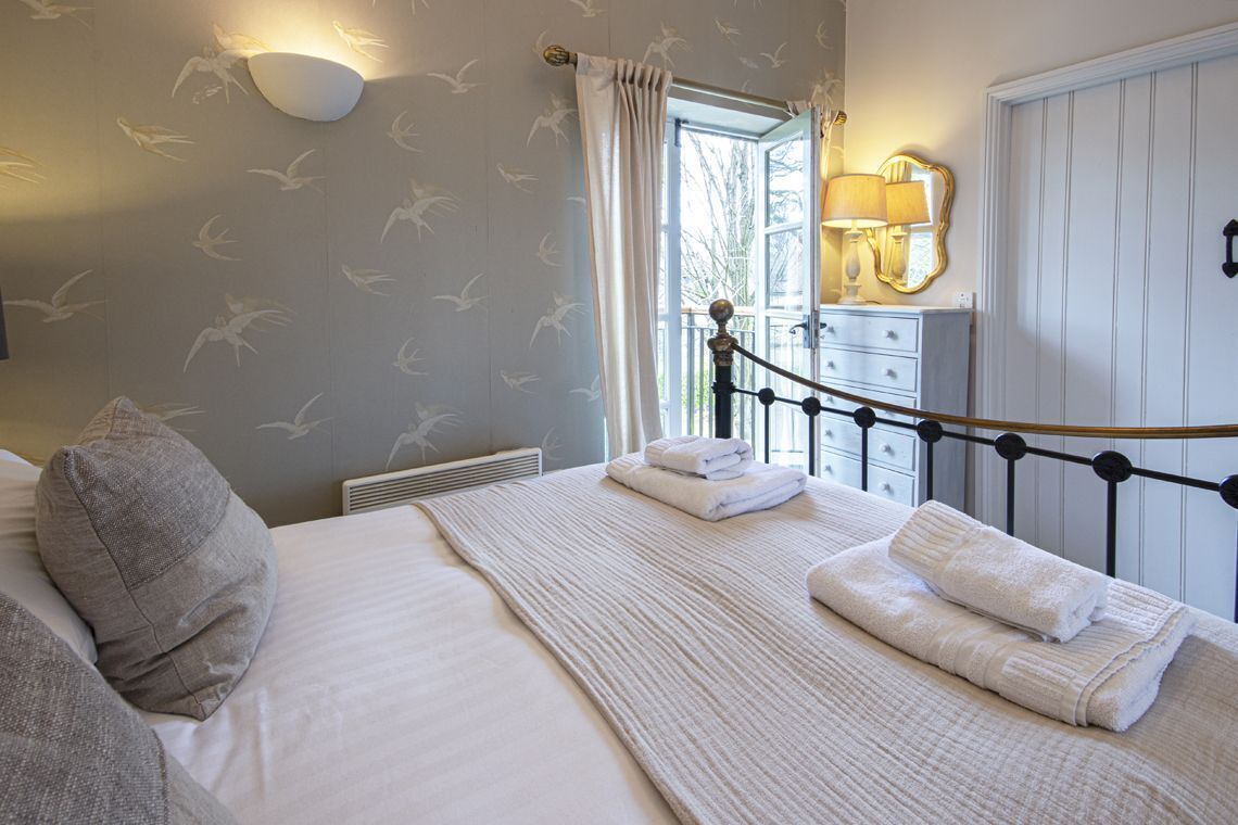 A neatly made bed with white linens and a gray pillow, a bedside table with a lamp, and a window with curtains.