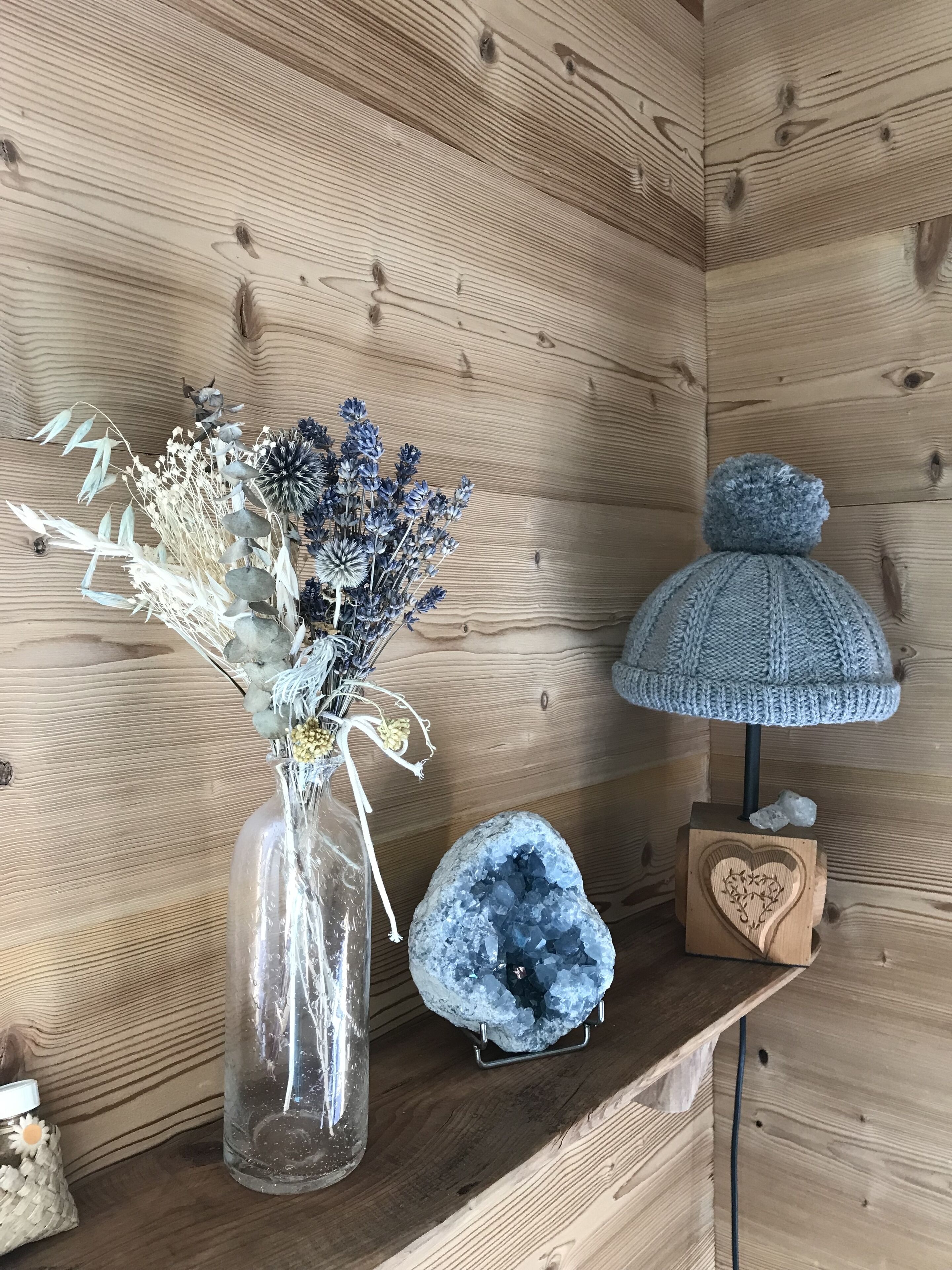 A wooden shelf with a vase of dried flowers, a crystal, and a knitted lamp.