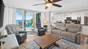 A modern living room with a sofa, armchair, and coffee table, a ceiling fan, and a view of the ocean through large sliding doors.