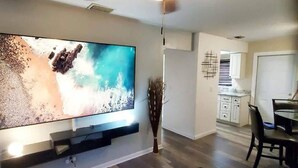 Ein modernes Wohnzimmer mit Flachbildfernseher an der Wand, einem Essbereich mit Tisch und Stühlen sowie einer Küche im Hintergrund.