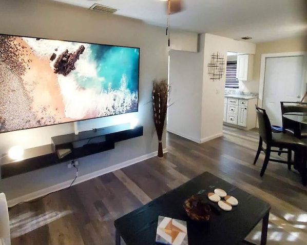 A modern living room with a flat-screen TV mounted on the wall, a dining area with a table and chairs, and a kitchen in the background.