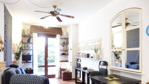 A living room with a sofa, a dining table with chairs, a ceiling fan, a bookshelf, and a door leading to a balcony. - STUDIO APARTMENT WITH SMALL GARDEN (Lido)