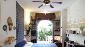 A bedroom with two beds, a ceiling fan, a bookshelf, and a view of the outdoors. - STUDIO APARTMENT WITH SMALL GARDEN (Lido)