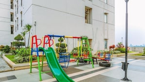 Une aire de jeux avec un toboggan vert et des équipements colorés, entourée d’un espace paysager agrémenté de plantes et d’un bâtiment en arrière-plan.