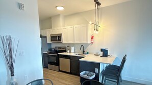 A modern kitchen with white cabinets, a microwave, and a sink. There is a dining area with a glass table and chairs, and a small plant on the table.
