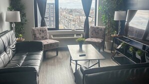 A modern living room with a leather sofa, two armchairs, a coffee table, and a TV. Large windows with curtains offer a city view.