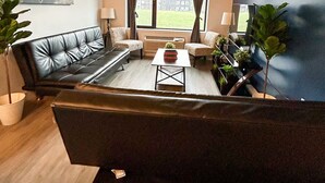 Ein modernes Wohnzimmer mit einer schwarzen Ledersofa, einem Couchtisch und zwei Sesseln. Es gibt ein großes Fenster mit Vorhängen, ein Bild an der Wand und eine Deckenleuchte.