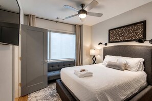 A hotel room with a bed, a ceiling fan, a TV, a window, and a wall-mounted lamp.